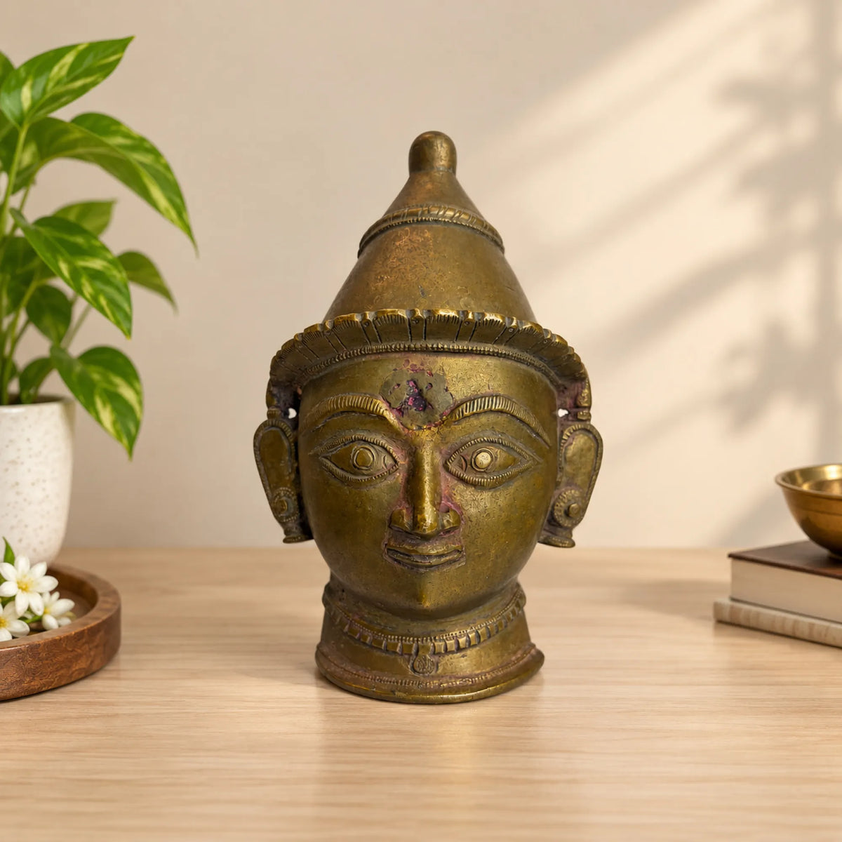 Antique brass Gauri devi face mask styled on a wooden console with a plant
